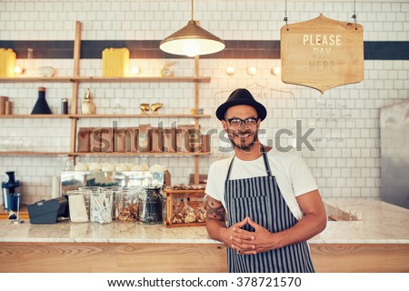 Portrait of happy young man wearing an apron and hat leaning to a cafe counter. Male barista standing at coffee shop looking at a camera and smiling.