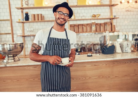 Portrait of happy young barista at work. Caucasian man wearing apron and hat standing in front of cafe counter with cup of coffee and looking at camera smiling.