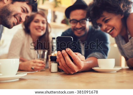 Group of friends having fun at the cafe and looking at smart phone. Man showing something to his friends sitting by, focus on mobile phone.
