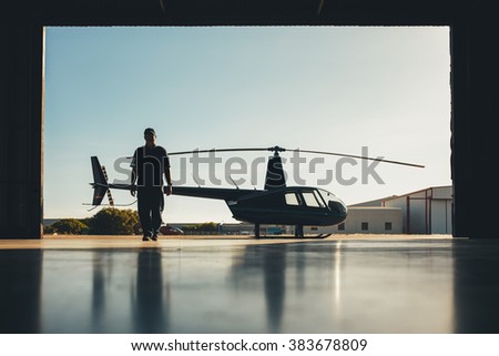 Silhouette of helicopter with a pilot in the airplane hangar. Pilot walking away from helicopter parked outside the hangar.