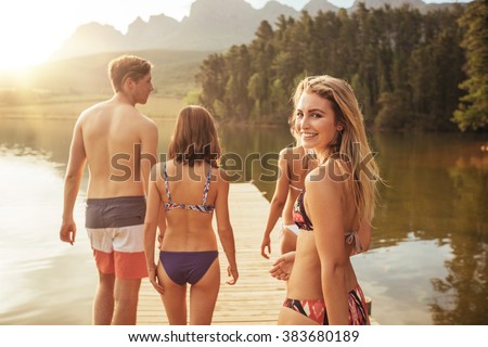 Portrait of beautiful young girl in bikini looking over her shoulder with her friends on pier. Group of young people walking together on a jetty at the lake on a sunny day.