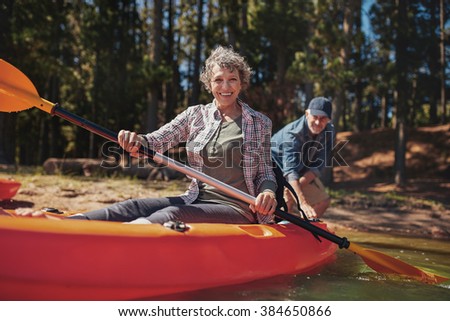 Portrait of happy senior woman in a kayak holding paddles. Woman canoeing with man in background on the lake.