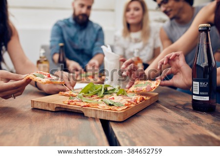 Close up shot of pizza on table, with group of young people sitting around and picking up a portion. Friends partying and eating pizza.