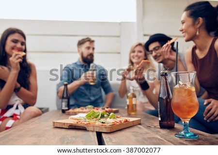 Group of multi-ethnic friends enjoying pizza and beer in party. Young people having a party. Focus on pizza and cocktail lying on wooden table.
