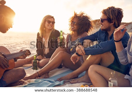 Portrait of group of young friends having a party on the beach in evening. Men and women drinking beers and listening to friend playing guitar.