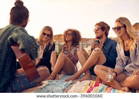 Group of young people listening to friend playing guitar outdoors. Diverse group of friends hanging out at beach. Young men and women drinking beers and enjoying music.