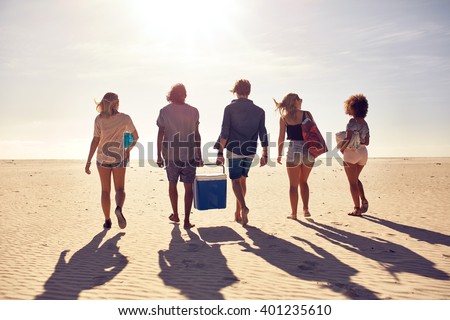 Rear view portrait of group of young people walking on the beach carrying a cooler box. Young men and woman on sea shore. Friends on beach vacation.