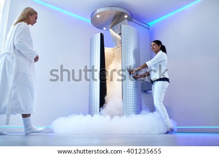Full length portrait of nurse opening the door of cryosauna, with woman in bathrobe at the clinic. Female going for cryotherapy treatment in freezing cabinet.