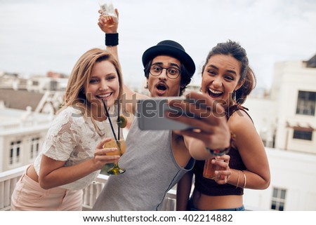 Excited young people taking self portrait with mobile phone during a party. Happy young man and woman taking self portrait at rooftop party. Multiracial people having fun in party with drinks.