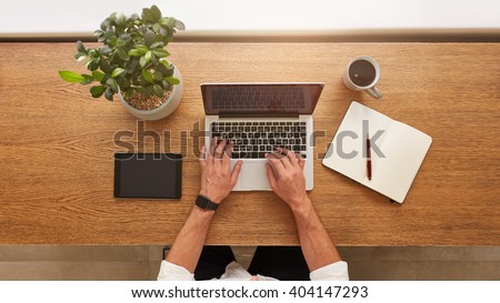 Directly above view of human hands typing on laptop. Laptop, digital tablet, diary, coffee cup and potted plant on work desk. Man working from home.