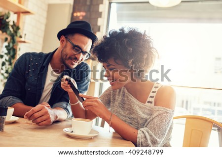 Portrait of happy young man and woman sitting together at a restaurant and looking at a mobile phone. Young friends looking at smart phone while sitting in cafe