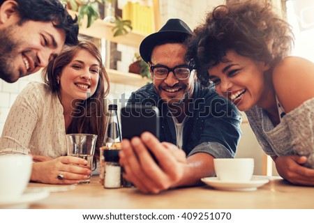 Group of friends sitting together in a cafe looking at mobile phone and smiling. Young guy showing something to his friends on his smart phone.