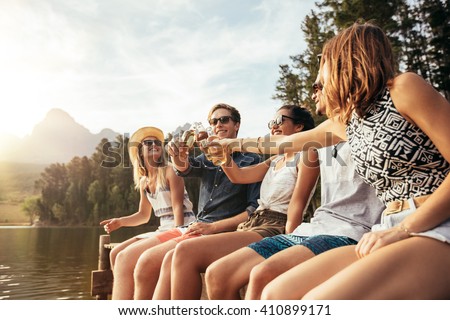 Portrait of young people sitting on a jetty and toasting beers on a sunny day. Happy young men and women hanging out at the lake.