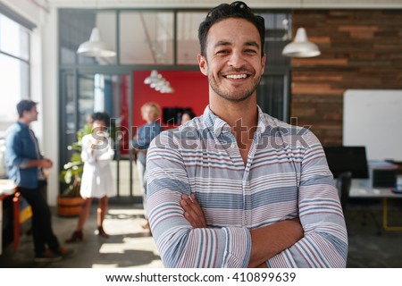 Portrait of a handsome young business man standing with arms crossed in an office. Smart caucasian young male designer with his colleagues in background.