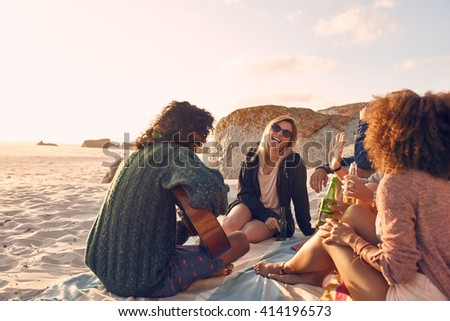 Portrait of young man playing guitar for friends. Group of friends having fun at the beach party. Young men and women sitting on the beach dinking beer.