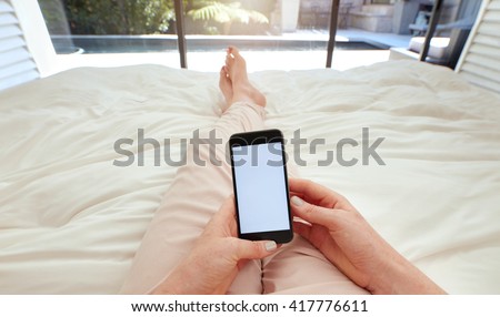 Closeup image of woman lying on a bed holding a smart phone with blank screen. POV shot of woman relaxing in bedroom using touch screen cellphone.