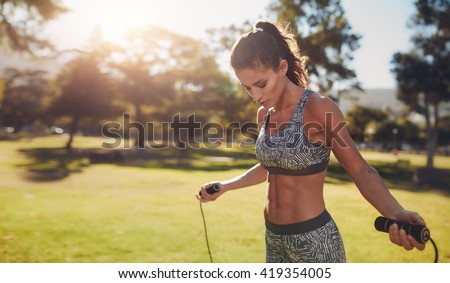 Horizontal shot of muscular woman with skipping rope outdoors in nature. Fitness female doing skipping workout with jump rope in a park on a sunny day.