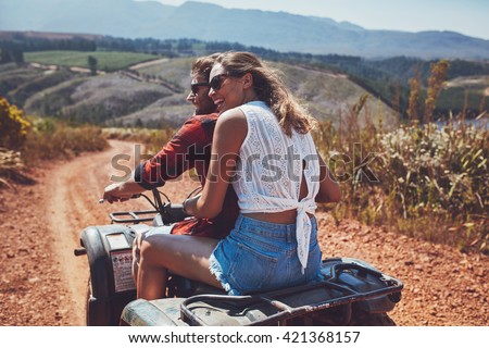 Rear view shot of young couple riding on a quad bike in countryside and looking away smiling. Woman sitting behind her boyfriend driving an quad on country road.