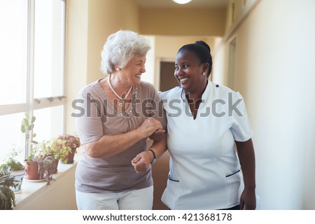 Portrait of smiling home caregiver and senior woman walking together through a corridor. Healthcare worker taking care of elderly woman.