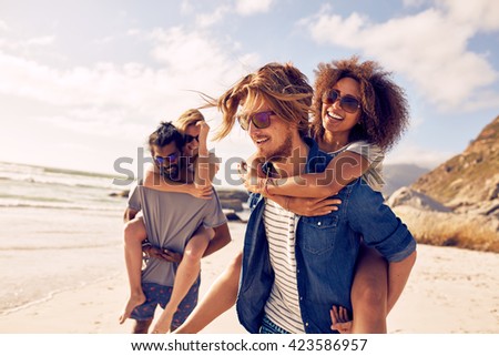 Two happy young men giving their girlfriends piggyback rides. Group of young people enjoying themselves during summertime at the beach.