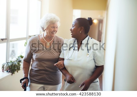 Senior woman walking in the nursing home supported by a caregiver. Nurse assisting senior woman.