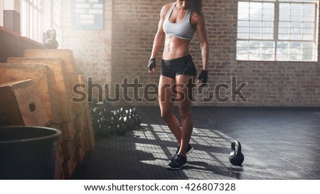Cropped shot of muscular woman standing in gym. Fitness female model in sportswear with kettle bell on floor.
