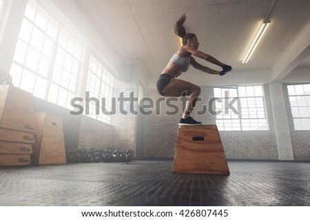 Side view image of fit young woman doing a box jump exercise. Muscular woman doing a box squat at the gym