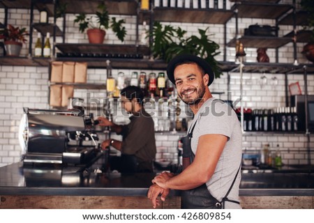 Portrait of happy young male coffee shop owner standing with barista working behind the counter making drinks.