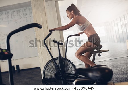 Fit young woman using exercise bike at the gym. Fitness female using air bike for cardio workout at crossfit gym.