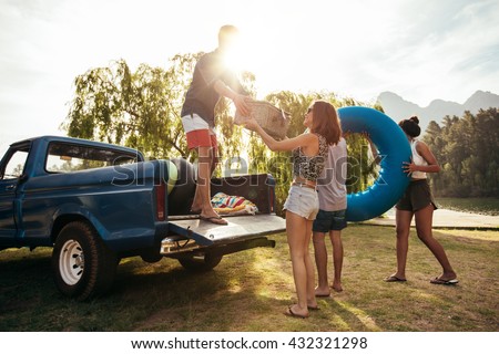 Group of young friends on picnic by the lake. Men and women unloading pickup truck on camping trip, carrying picnic basket and inflatable tube on sunny day.