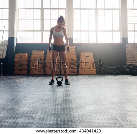 Full length shot of fit and strong woman standing in crossfit gym. Tough fitness female model with kettle bell on floor.