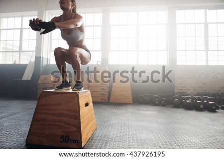 Shot of a young woman jumping onto a box as part of exercise routine. Fitness woman doing box jump workout at crossfit gym.