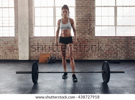 Full length portrait of fit young woman standing at gym with barbells on floor. Tough crossfit female at gym.