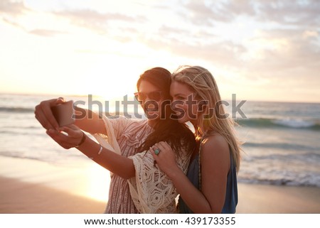 Happy young women enjoying vacation together having fun on the beach and taking selfie photo using smartphone camera.