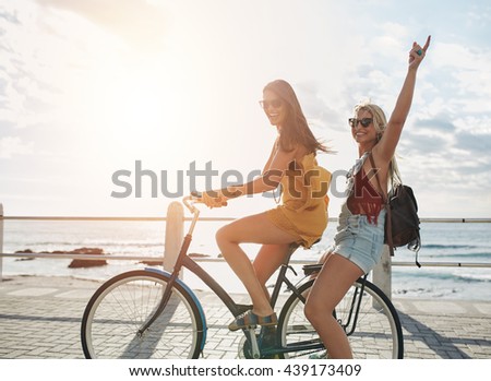 Outdoor shot of happy young women enjoying bike ride. Two female friends on a bicycle at seaside promenade on a sunny day.