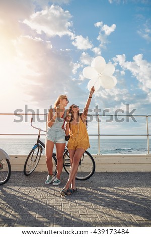 Outdoor shot of two women standing together with bicycles and balloons on seaside road. Best friends having fun on their vacation holding balloons and eating icecream.