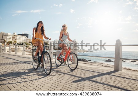 Two young female friends riding their bicycles on the seaside promenade. Cheerful young women riding bikes at the waterfront on a summer day.