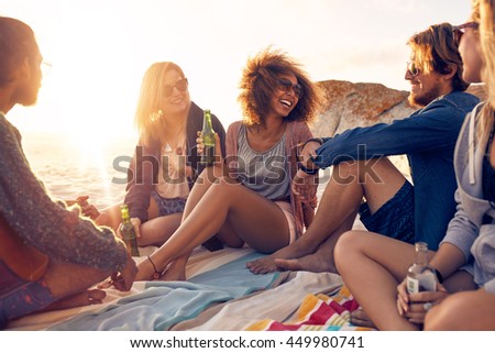 Mixed race friends having fun at the beach. Group of happy young people sitting together at the beach talking and drinking beers.