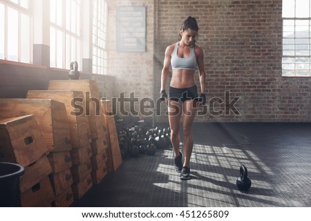 Full length shot of young woman in sportswear taking a walk in the cross fit gym and looking at kettle bell on floor. Fitness female getting ready for intense crossfit workout.
