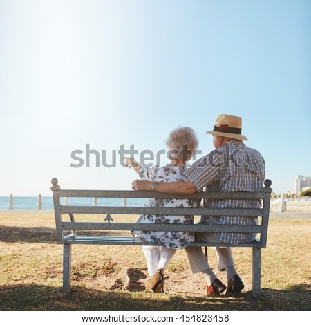 Rear view of senior couple relaxing on a bench with woman pointing out to sea. Retired man and woman sitting on a bench outdoors and enjoying the view.
