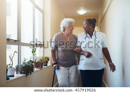 Portrait of smiling healthcare worker walking and talking with senior woman. Happy elder woman gets help from nurse for a walk through nursing home.