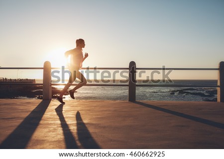 Side view of fitness woman running on a road by the sea. Sportswoman training on seaside promenade at sunset.