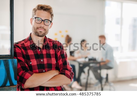 Portrait of a positive looking young business professional standing with his arms crossed with coworkers talking in the background.