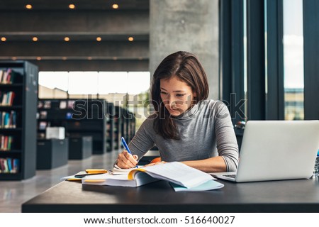 Female student taking notes from a book at library. Young asian woman sitting at table doing assignments in college library.