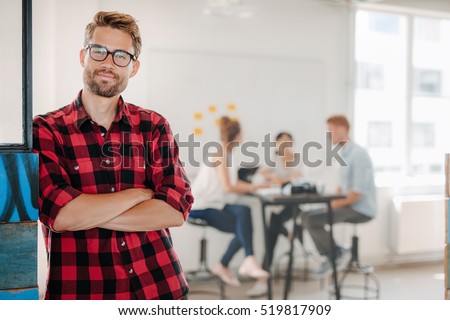 Portrait of relaxed young man standing in office with colleagues meeting in background.