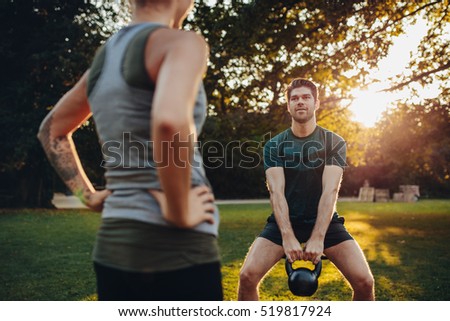 Young man doing weight training with personal female trainer in the park. Caucasian male exercising with kettlebell.