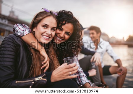 Shot of young female friends having fun with drinks on the lake. Group of people enjoying a day by the lake.