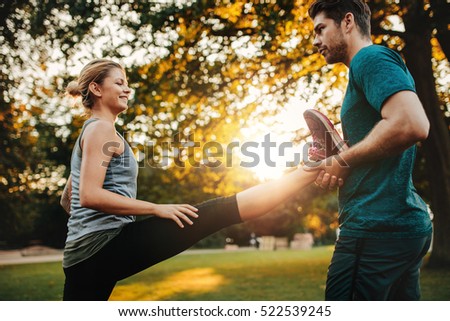 Personal trainer holding leg of woman stretching in park. Female exercising with support from her coach.