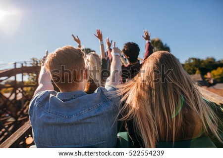 Rear view shot of young people on a thrilling roller coaster ride at amusement park. Group of friends having fun at fair and enjoying on a ride.