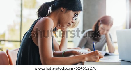 Close up shot of young woman writing notes with classmates studying in background. Students learning in college library.